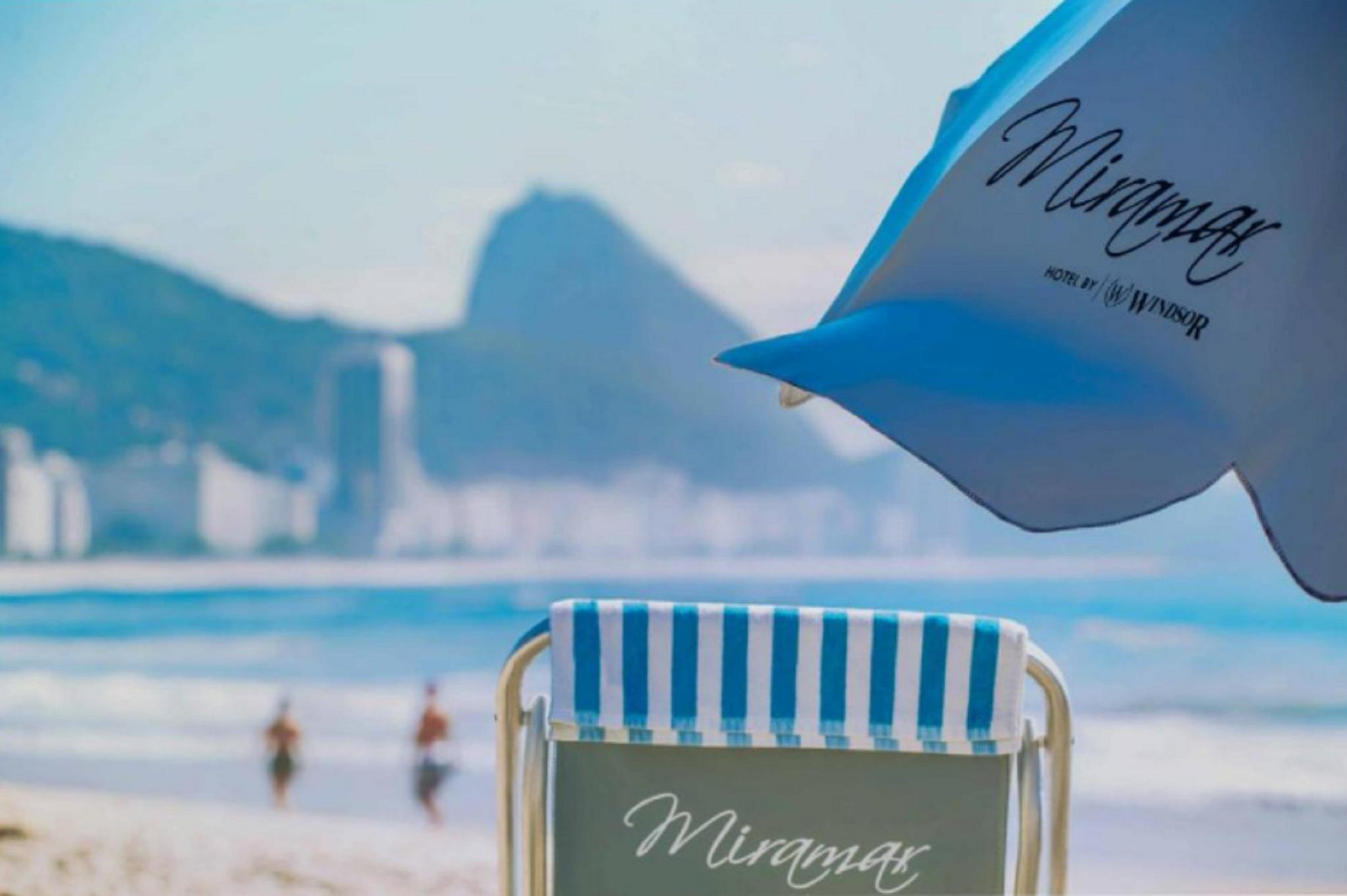 Miramar By Windsor Copacabana Río de Janeiro Exterior foto The photo features a beach scene with a lounge chair in the foreground. The chair has a striped blue and white design and displays the name "Miramar." In the background, there are blurred figures walking along the beach, with a hazy view of mountains